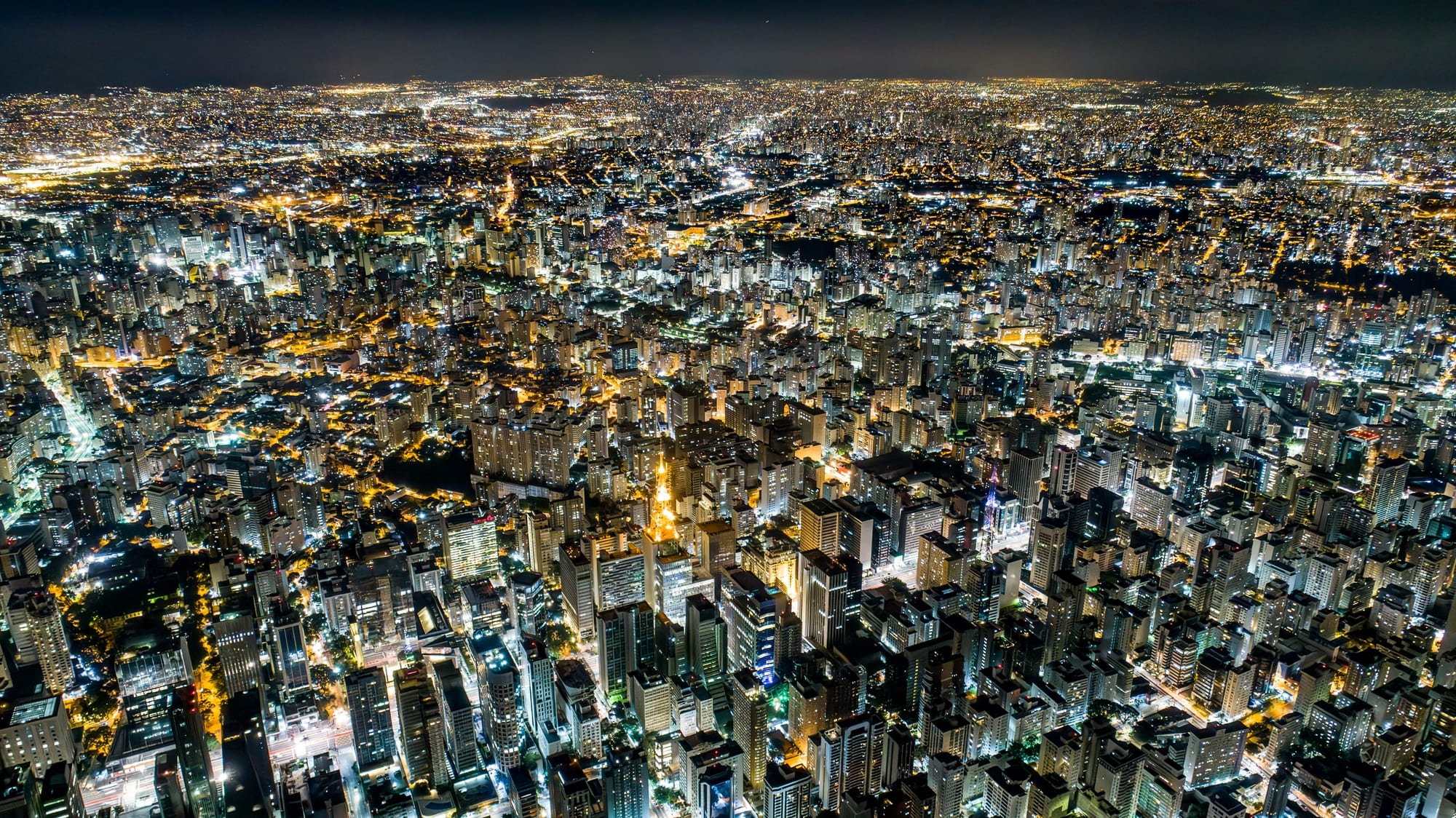 Bacurau Noturno 2, fotografia fine art de Bruno Boni. Vista aérea de São Paulo à noite, com a malha de avenidas iluminadas correndo em direção a um horizonte que não…
