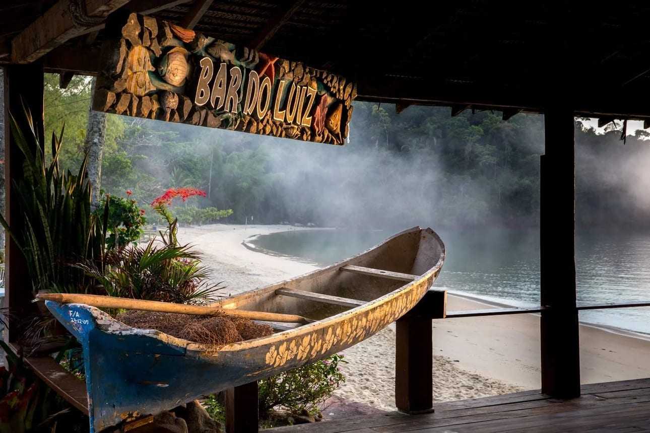 Bar do Luiz, fotografia fine art de Bruno Boni. Luz da manhã no Bar do Luiz, onde uma canoa de tronco de guapuruvu, típica da cultura caiçara, enfeita o bar. A enseada…