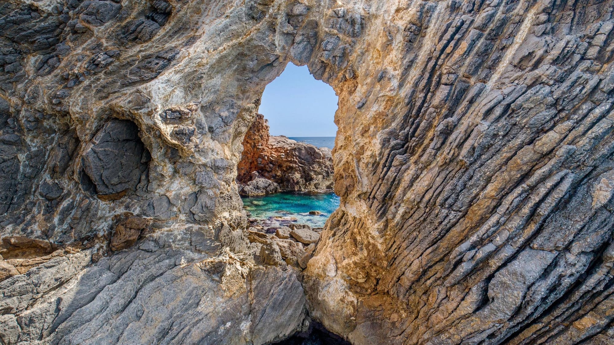 Cala Jondal Portal 1, fine art photograph by Bruno Boni. A natural rock arch in Cala Jondal, Ibiza, forms a perfect frame that reveals the crystalline waters of the cove…