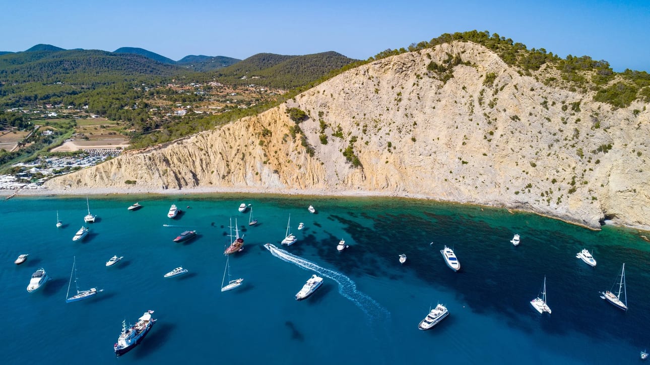 Cala Jondal, fotografia fine art de Bruno Boni. Vista aérea de Cala Jondal, Ibiza, mostrando a enseada preenchida por barcos e iates ancorados em águas cristalinas. O…