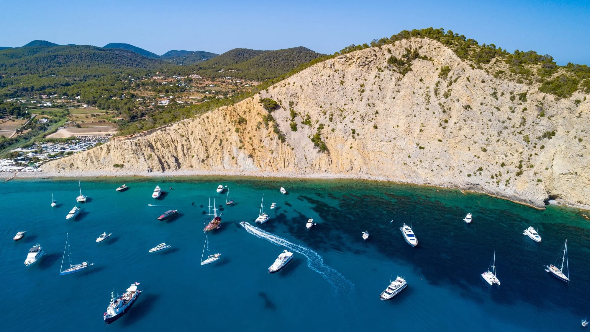 Cala Jondal, fotografia fine art de Bruno Boni. Vista aérea de Cala Jondal, Ibiza, mostrando a enseada preenchida por barcos e iates ancorados em águas cristalinas. O…