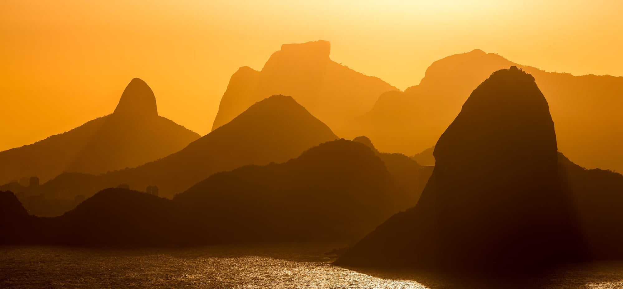 Camadas Cariocas, fotografia fine art de Bruno Boni. Silhuetas sobrepostas do relevo icônico do Rio sob a contraluz dourada do entardecer, destacando as formas familiares…