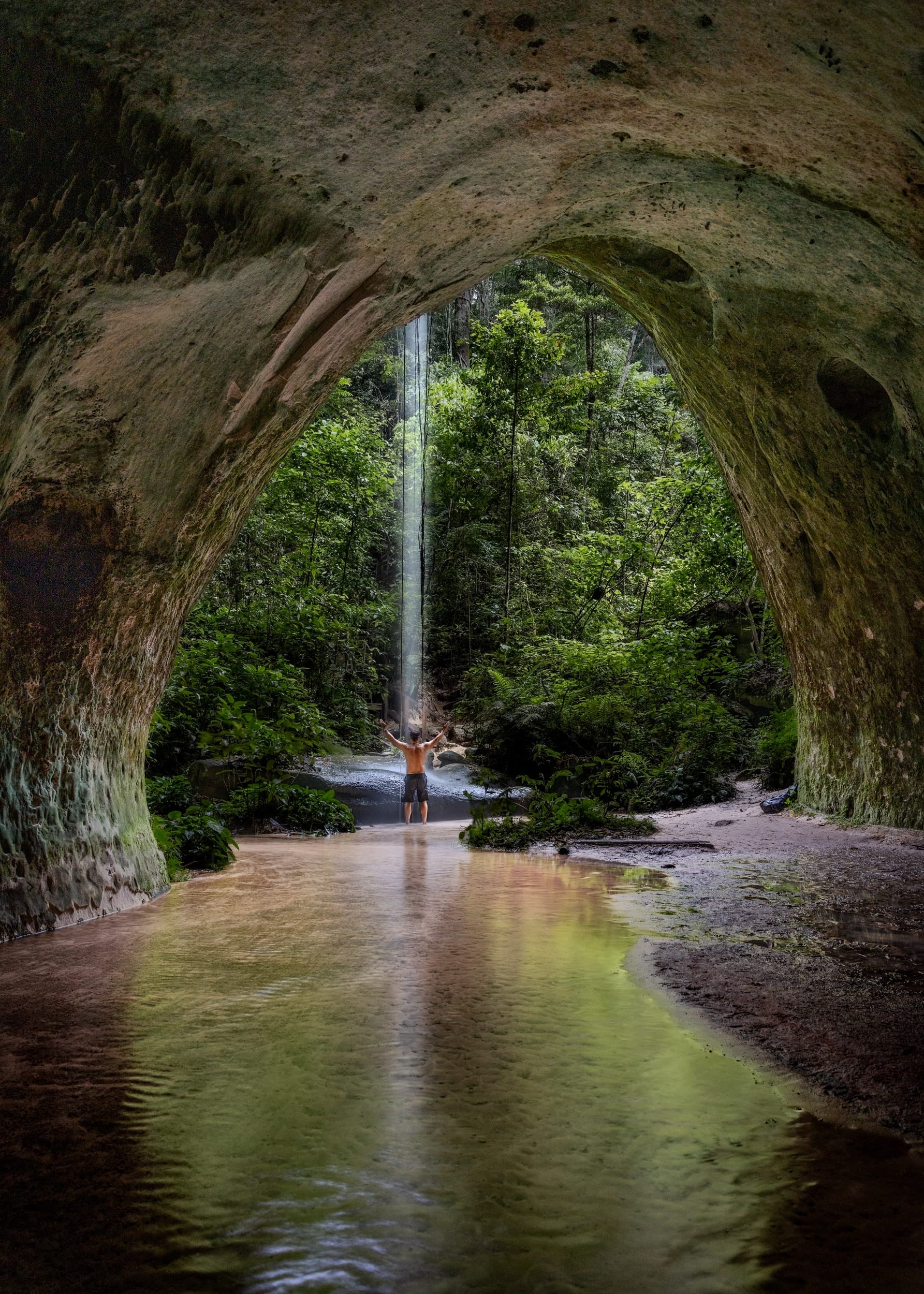 Caverna do Maroaga 2, fotografia fine art de Bruno Boni. Do interior da Caverna do Maroaga, o arco de arenito emoldura a floresta e o fio de água que desce da clareira. Uma…