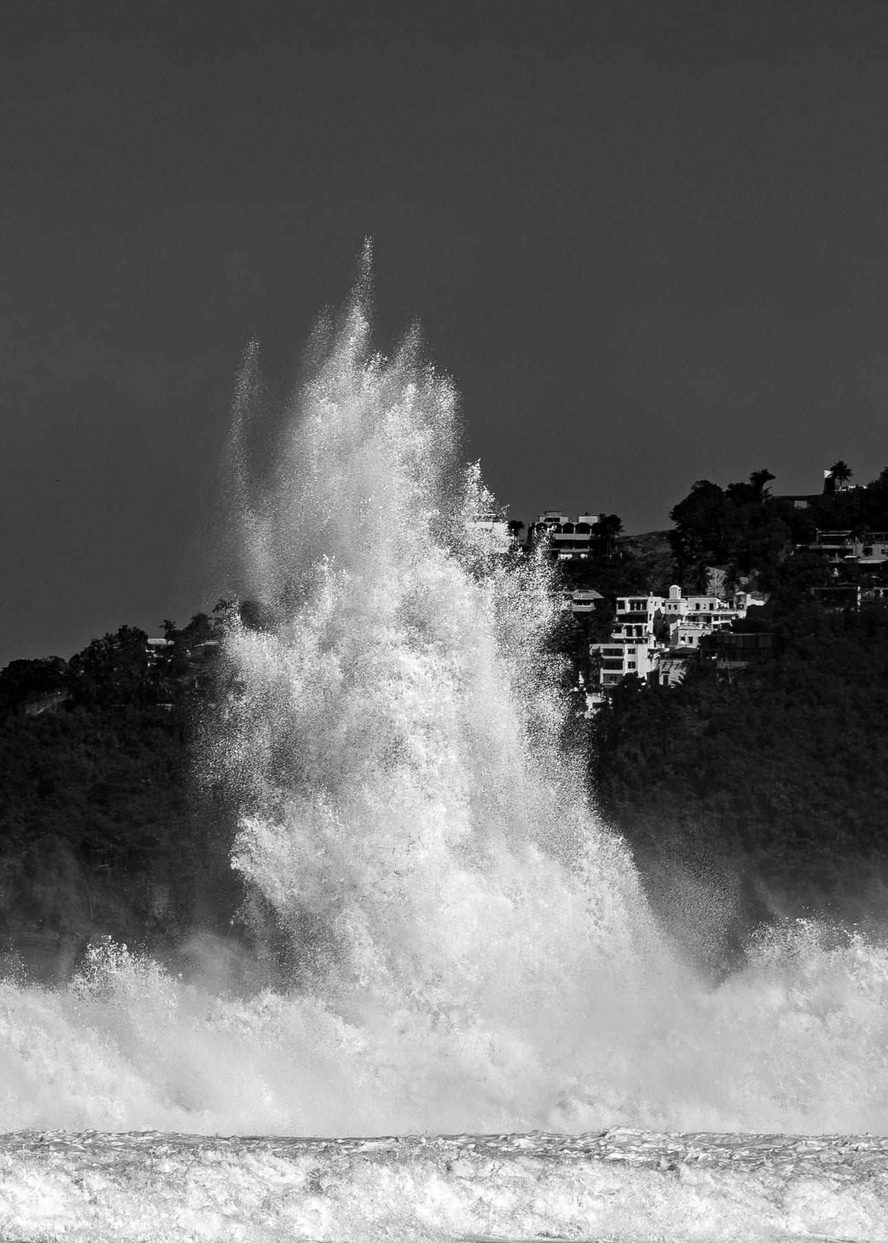 Encontro Explosivo 7, fotografia fine art de Bruno Boni. Uma coluna de água sobe vertical como um gêiser, ultrapassando em altura as casas na encosta de São Conrado, no Rio de…