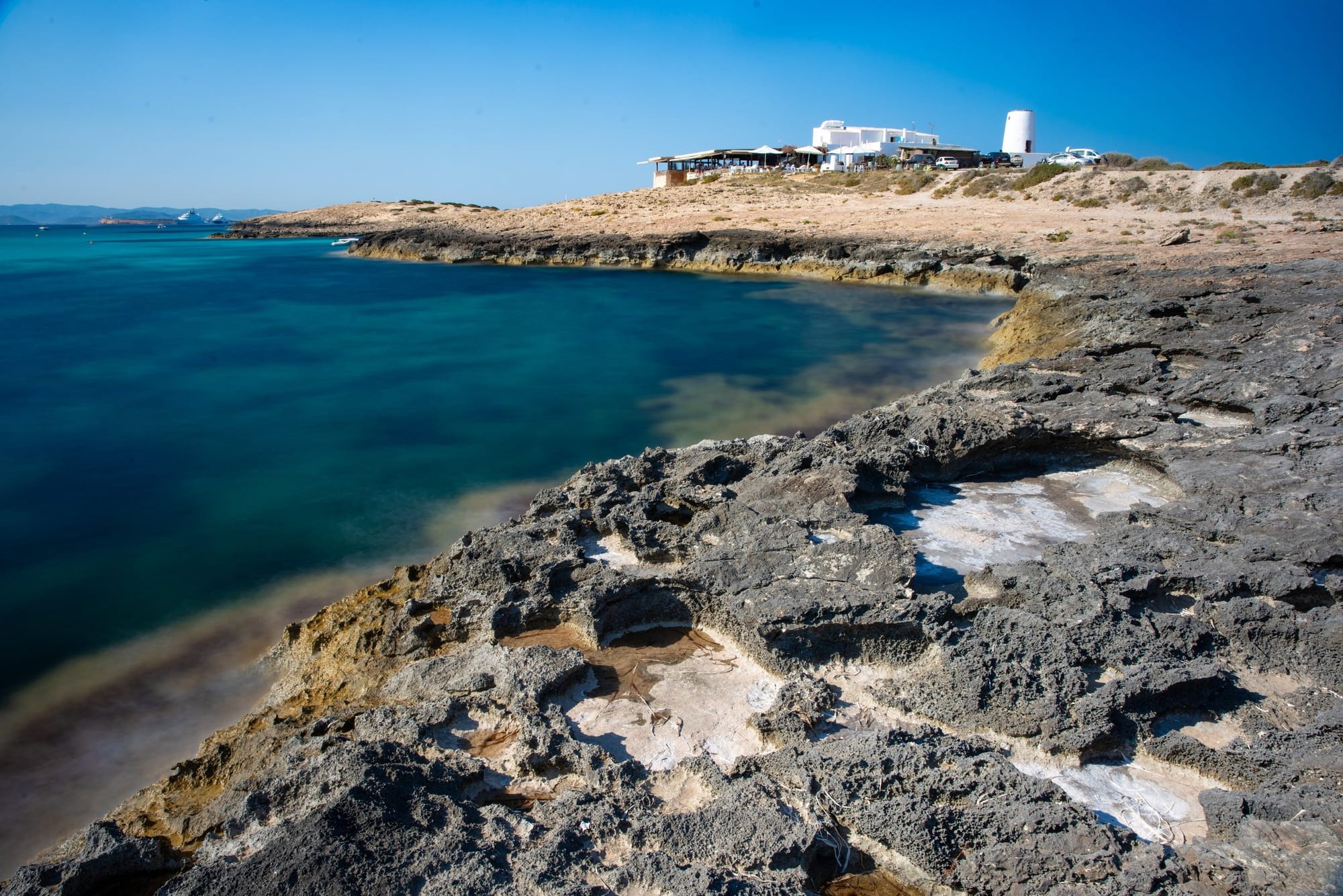 Es Molí: Do Moinho à Mesa, fotografia fine art de Bruno Boni. Vista de Es Molí de Sal em Formentera, revelando a costa rochosa que se curva suavemente até o emblemático restaurante…