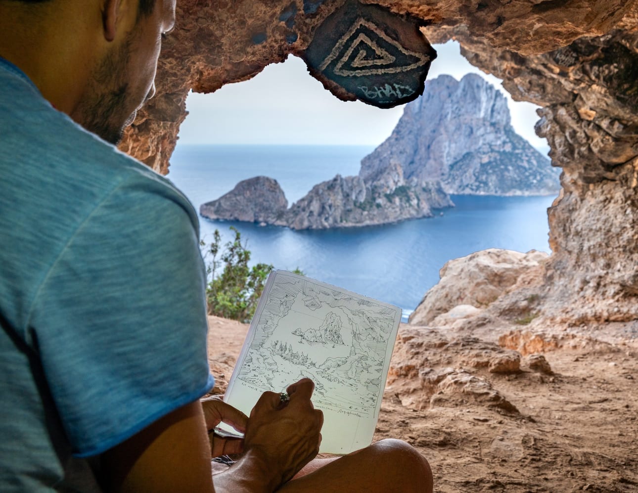 Portal de Es Vedrà - Meta-Arte, fotografia fine art de Bruno Boni. Um artista desenha Es Vedrà em seu caderno, enquanto o próprio ilhéu emerge colossal através do portal rochoso da…