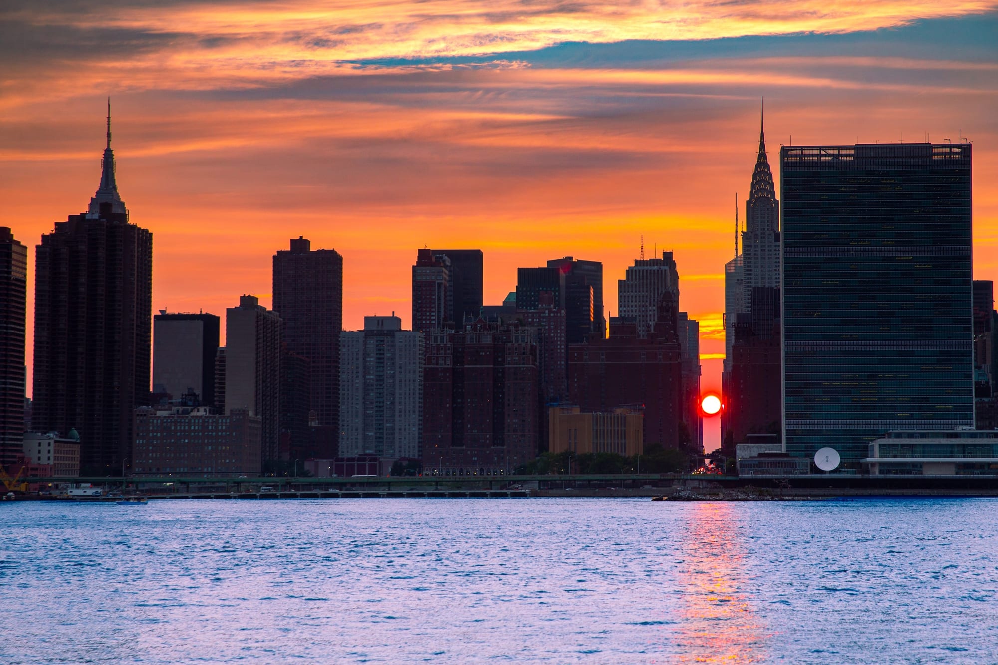 Manhattanhenge 2, fotografia fine art de Bruno Boni. O Manhattanhenge visto do outro lado do East River: o sol, reduzido a um ponto vermelho, atravessa a ilha inteira pelo…
