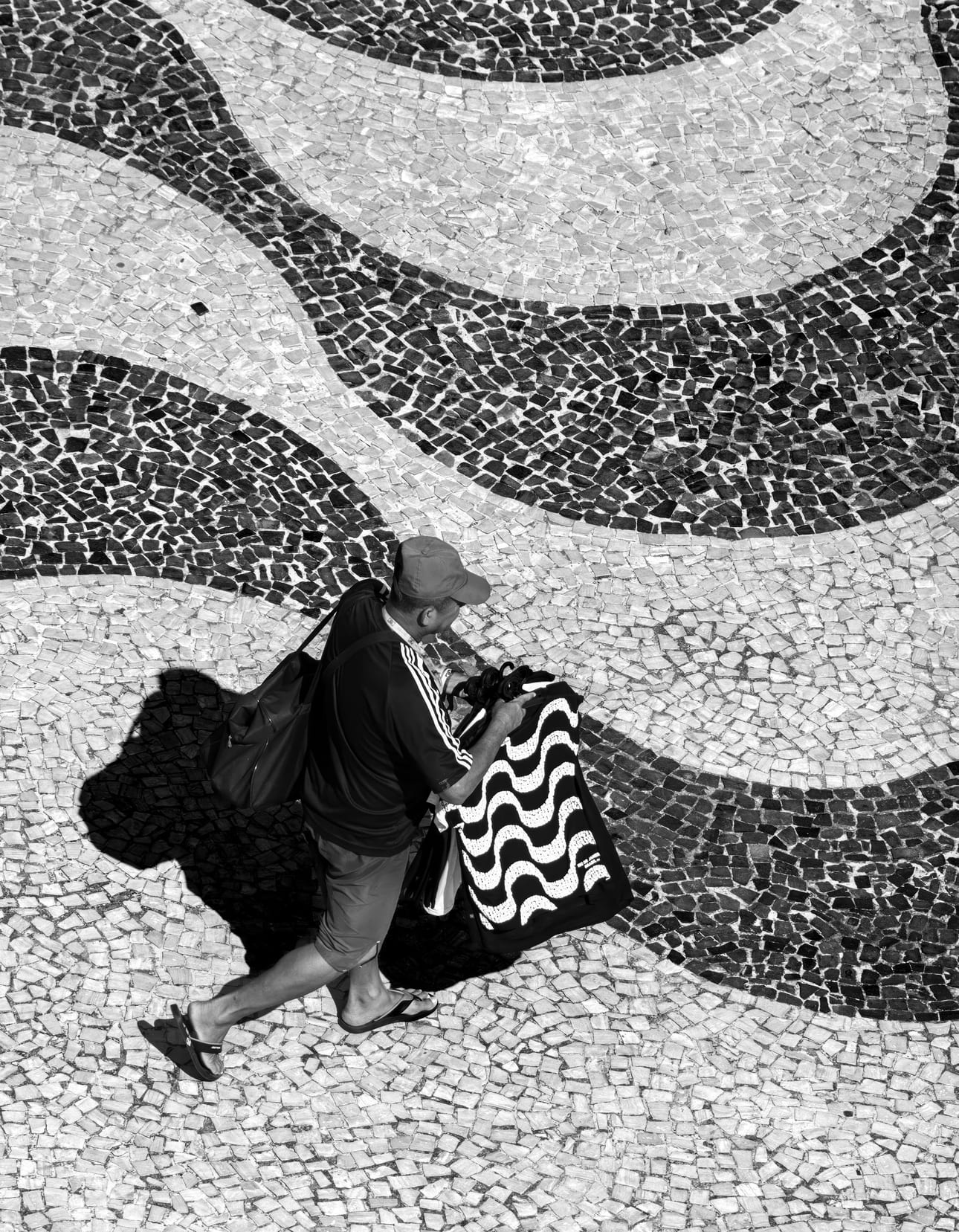 Meta Ondas, fotografia fine art de Bruno Boni. Ondas sobre ondas: a orla de São Conrado vira um mar de pedra, e nele caminha um único homem, vendendo camisetas que…
