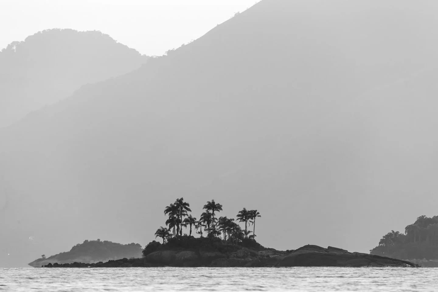 Pequena São João P&B, fine art photograph by Bruno Boni. Pequena São João rises as a silhouette before the imposing presence of Ilha Grande in the background, reinforcing the…